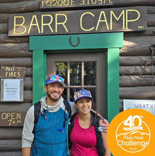 Two hikers in front of the lodge at Barr Camp, with a 40th Anniversary logo for Pikes Peak Challenge in the bottom corner.
