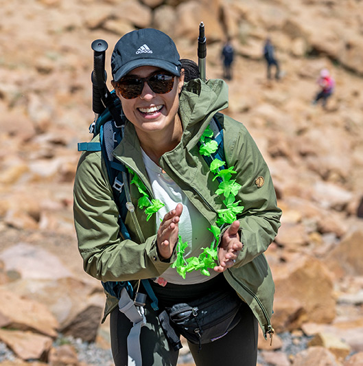 Woman wearing a hat, in hiking gear, clapping and smiling as she reaches the summit of Pikes Peak.